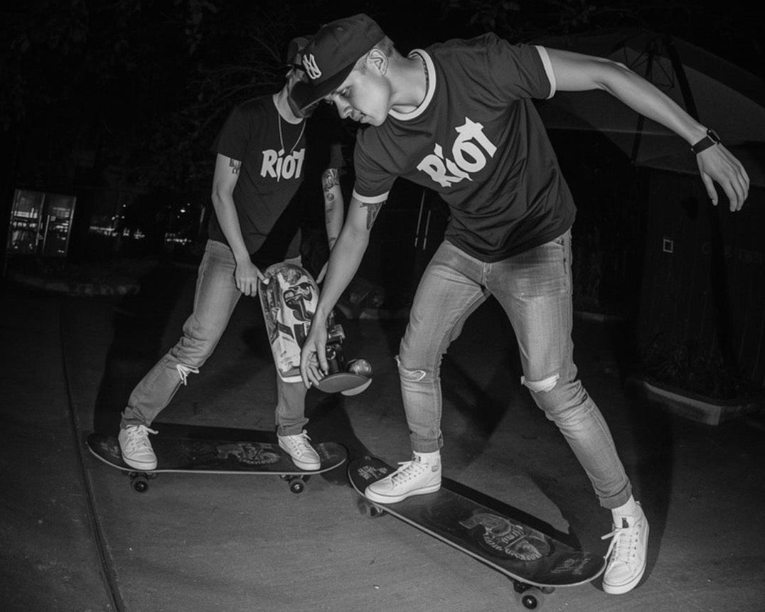 Two individuals wearing 'Riot' t-shirts with skateboards on a dark background
